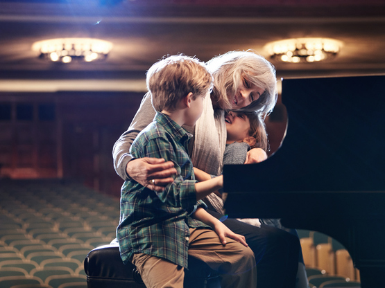 Zenész nagymama az unokáit öleli a zongoránál ülve.